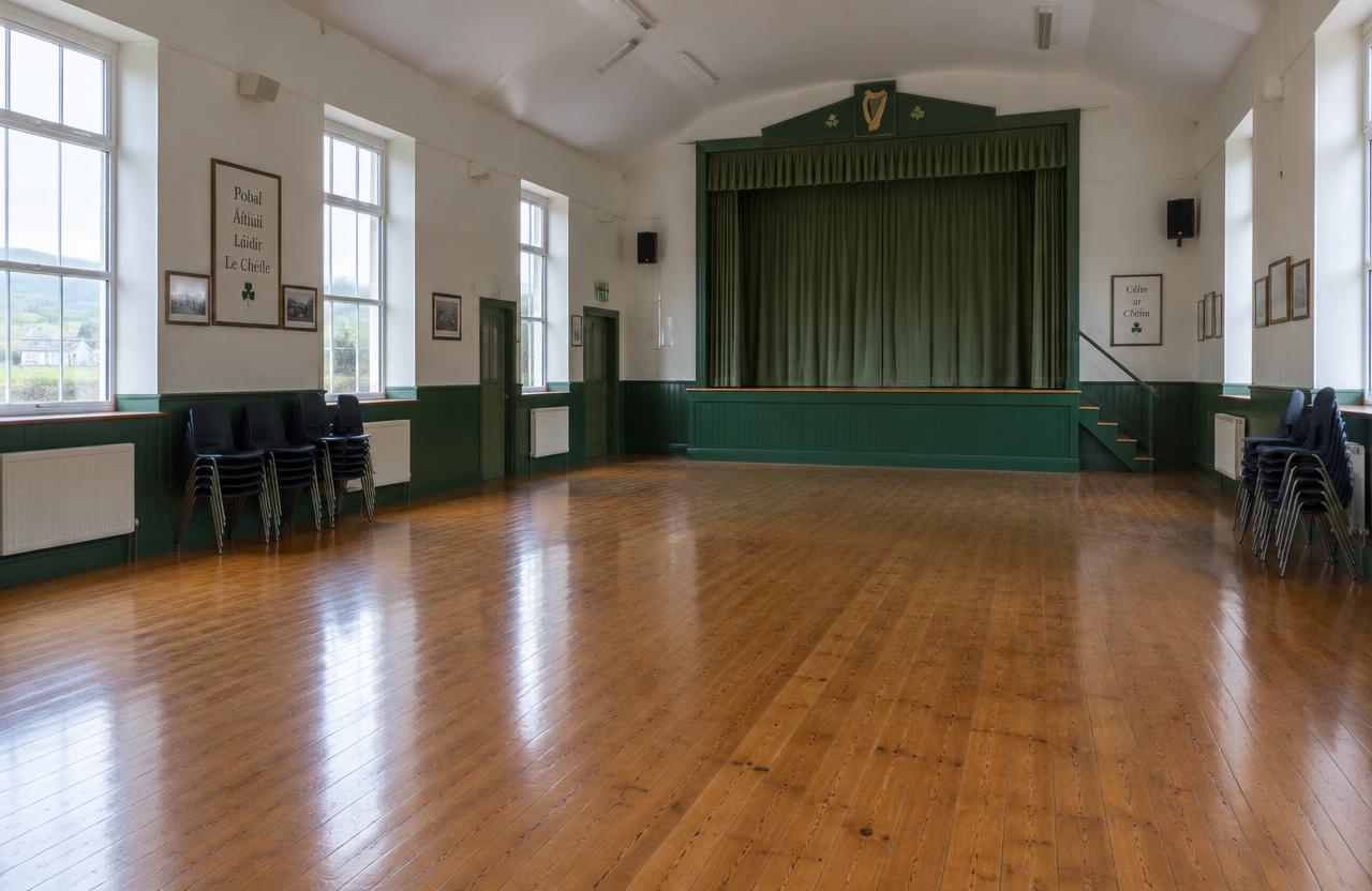 Inside the hall: a long timber floor, green panelling along the walls, stacked chairs and a stage with green curtains at the far end.